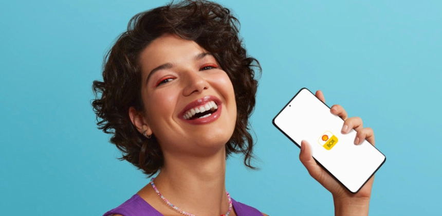 Mulher em um fundo azul, sorrindo e segurando um celular com o app Shell Box na tela